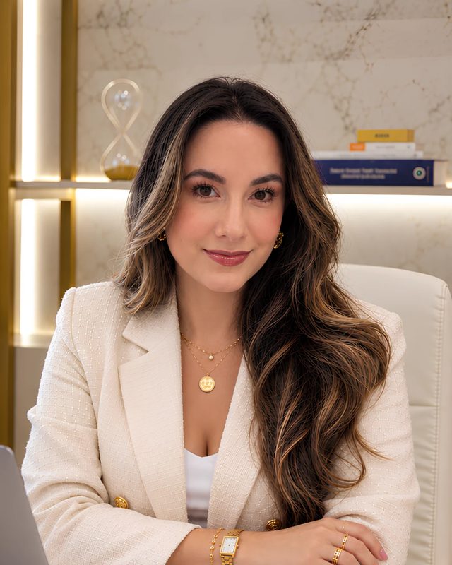 Allana Lisboa, psicóloga, sorrindo no consultório, de blazer creme.
